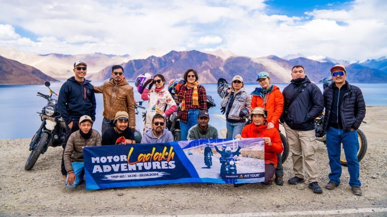 Nhóm du khách Fit Tour tạo dáng chụp ảnh tại bờ hồ Pangong, nơi cảnh quan hùng vĩ của dãy Himalaya phản chiếu trên mặt nước trong xanh