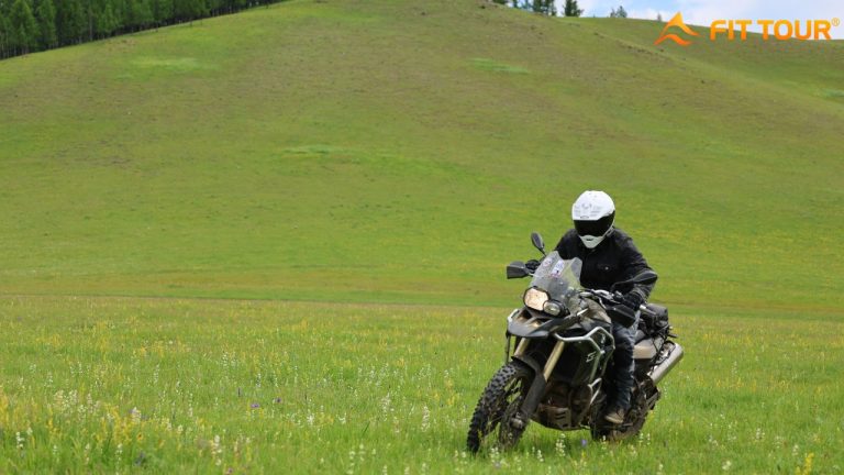 Người lái xe mô tô BMW GS đang chạy qua vùng thảo nguyên hoang sơ ở Mông Cổ, phía sau là đồi cỏ xanh và rừng thông thấp thoáng