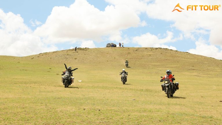 Các tay lái mô tô đang đổ dốc từ đỉnh đồi hoang sơ ở Mông Cổ, phía xa là nhóm hỗ trợ và ô tô địa hình đỗ trên đỉnh