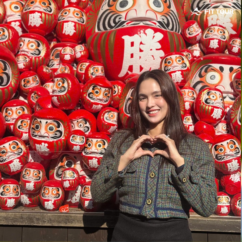 Khách hàng nữ rạng rỡ tạo dáng trái tim trước rừng búp bê Daruma đỏ rực, một điểm check-in "hot" trong tour.